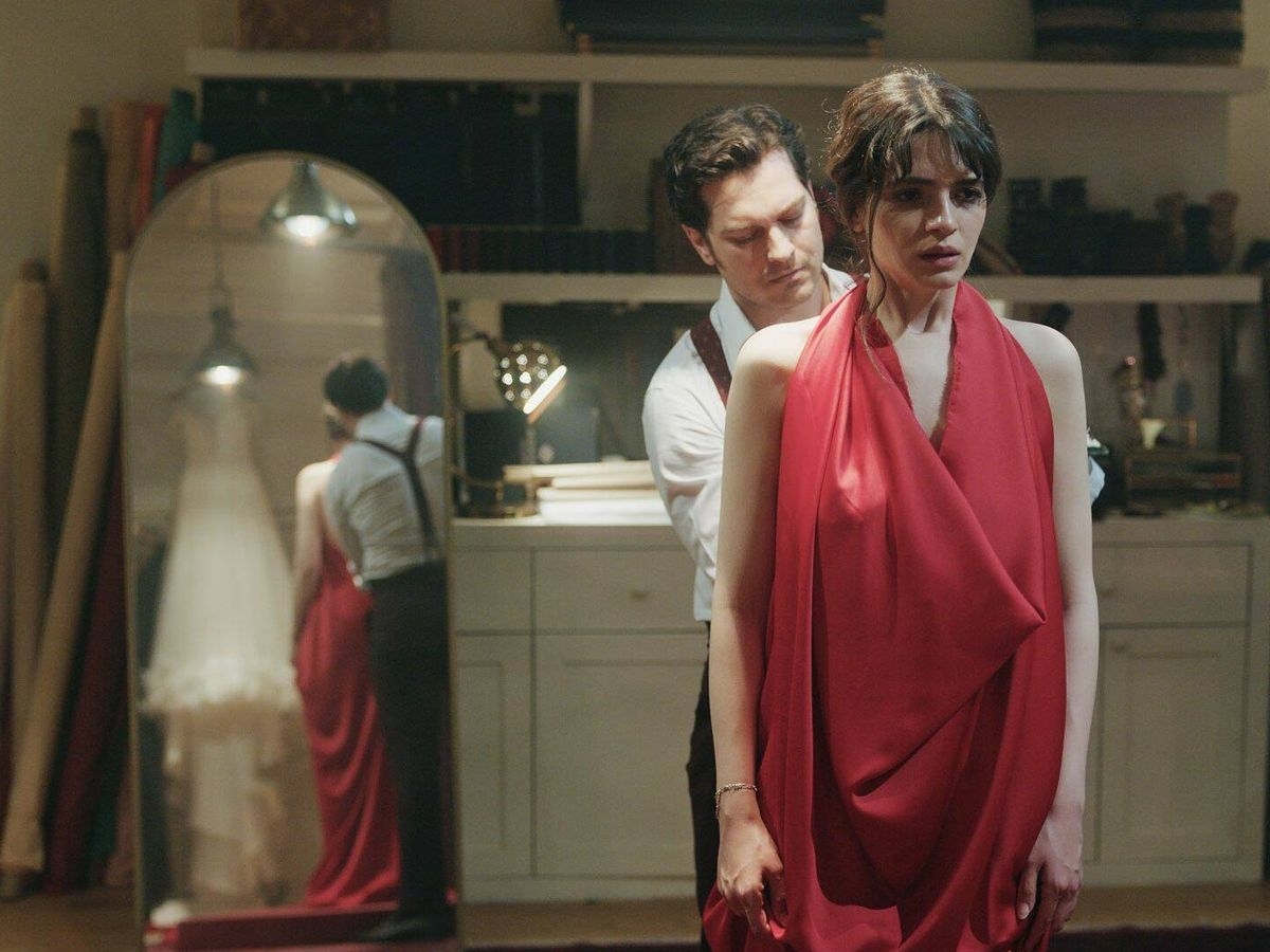 Un hombre arregla un vestido rojo drapeado en una mujer joven, ambos de pie en un taller con un espejo que muestra el reflejo de una vestido blanco en el fondo