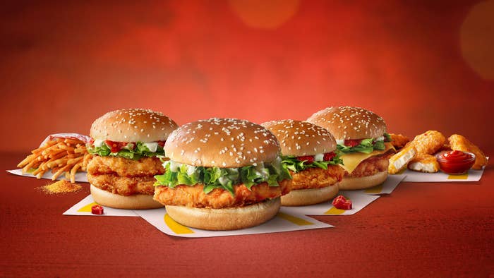 Four crispy chicken sandwiches with lettuce and sauce are displayed on paper mats; fries and chicken wings are in the background