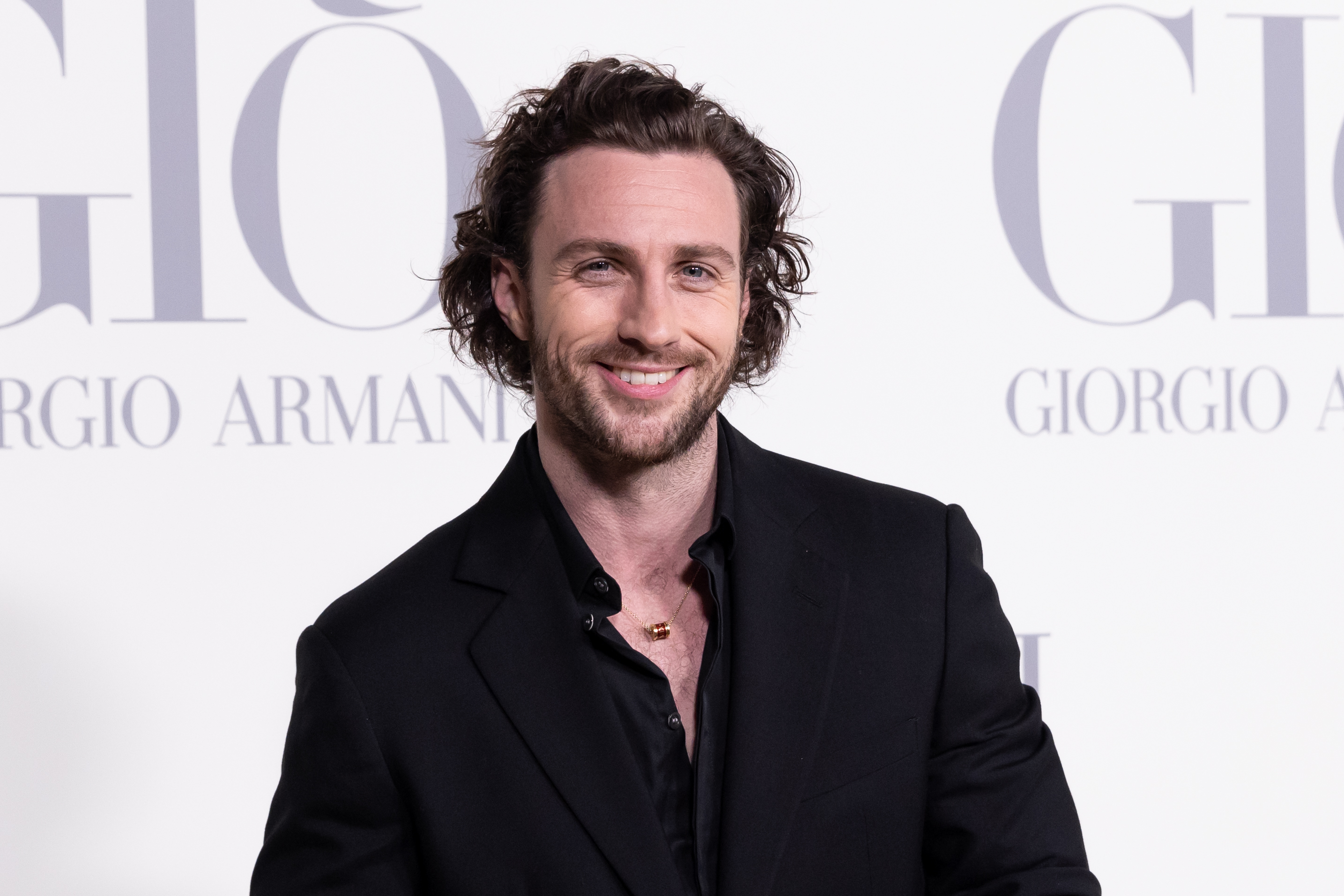 Aaron Taylor-Johnson at a Giorgio Armani event, wearing a black suit with a black shirt, smiling at the camera
