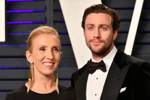 Sam Taylor-Johnson and Aaron Taylor-Johnson pose on the red carpet. Sam is wearing a black dress with gold earrings, and Aaron is in a black tuxedo with a bow tie