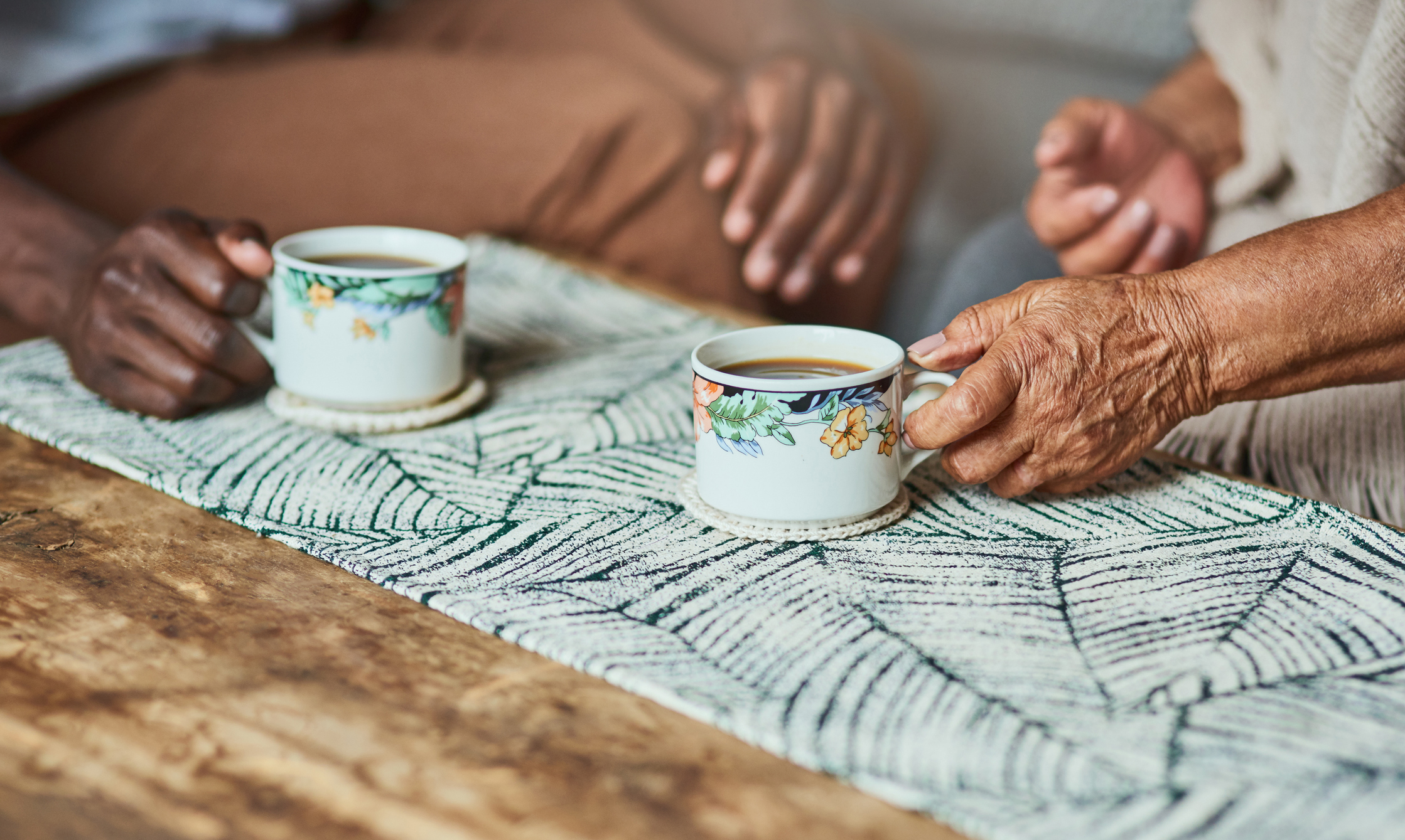 Two people sit at a table, each holding a cup of tea. The setting is casual with a patterned tablecloth and wooden table. Their faces are not visible