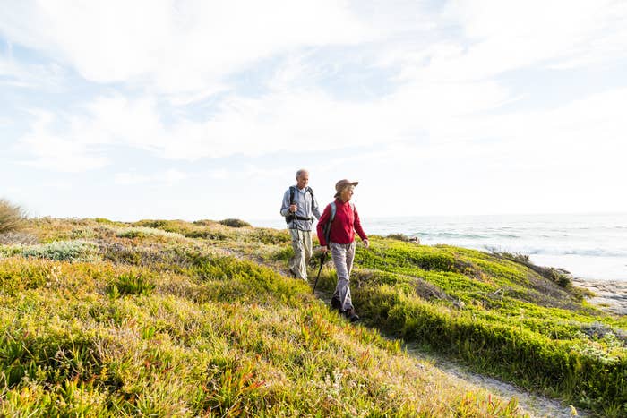 Two people are hiking on a grassy, coastal trail with the ocean in the background. They are wearing casual outdoor clothing and carrying backpacks