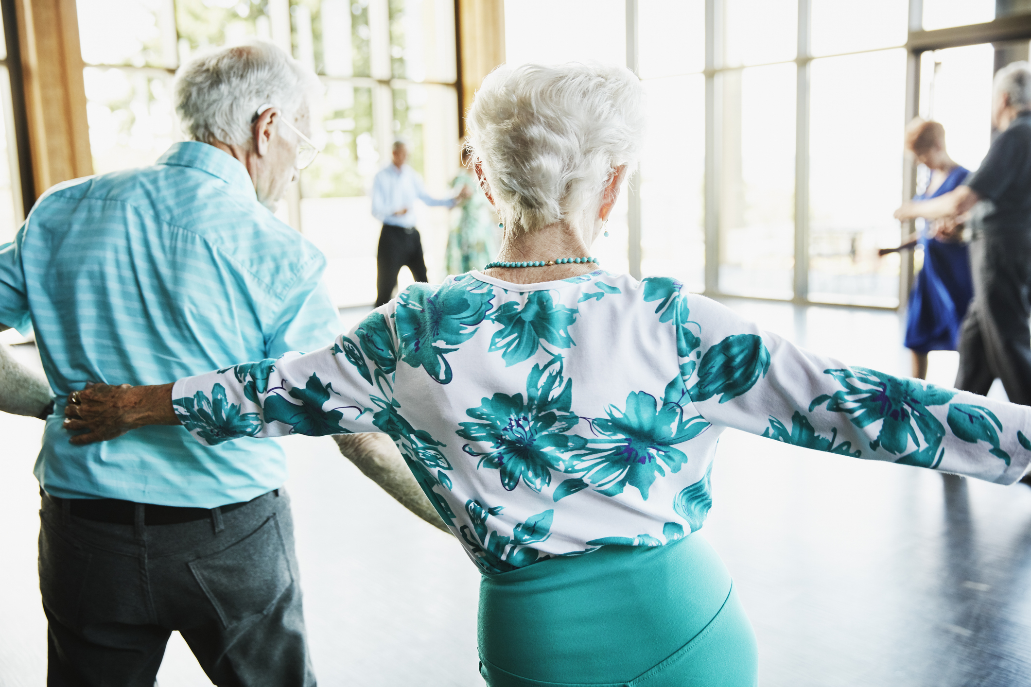 Elderly couple, backs to camera, dancing in a bright room; the woman in a floral top, the man in a light shirt. Other couples dance in the background
