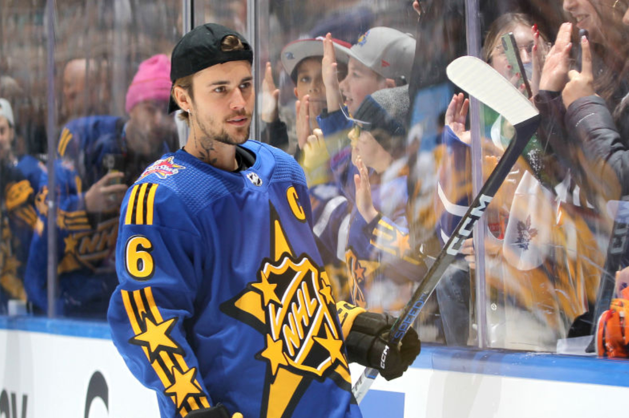 A hockey player with a beard and a backwards cap wears a blue "NHL All-Star" jersey with the number 6. He skates near a glass wall with cheering fans behind it