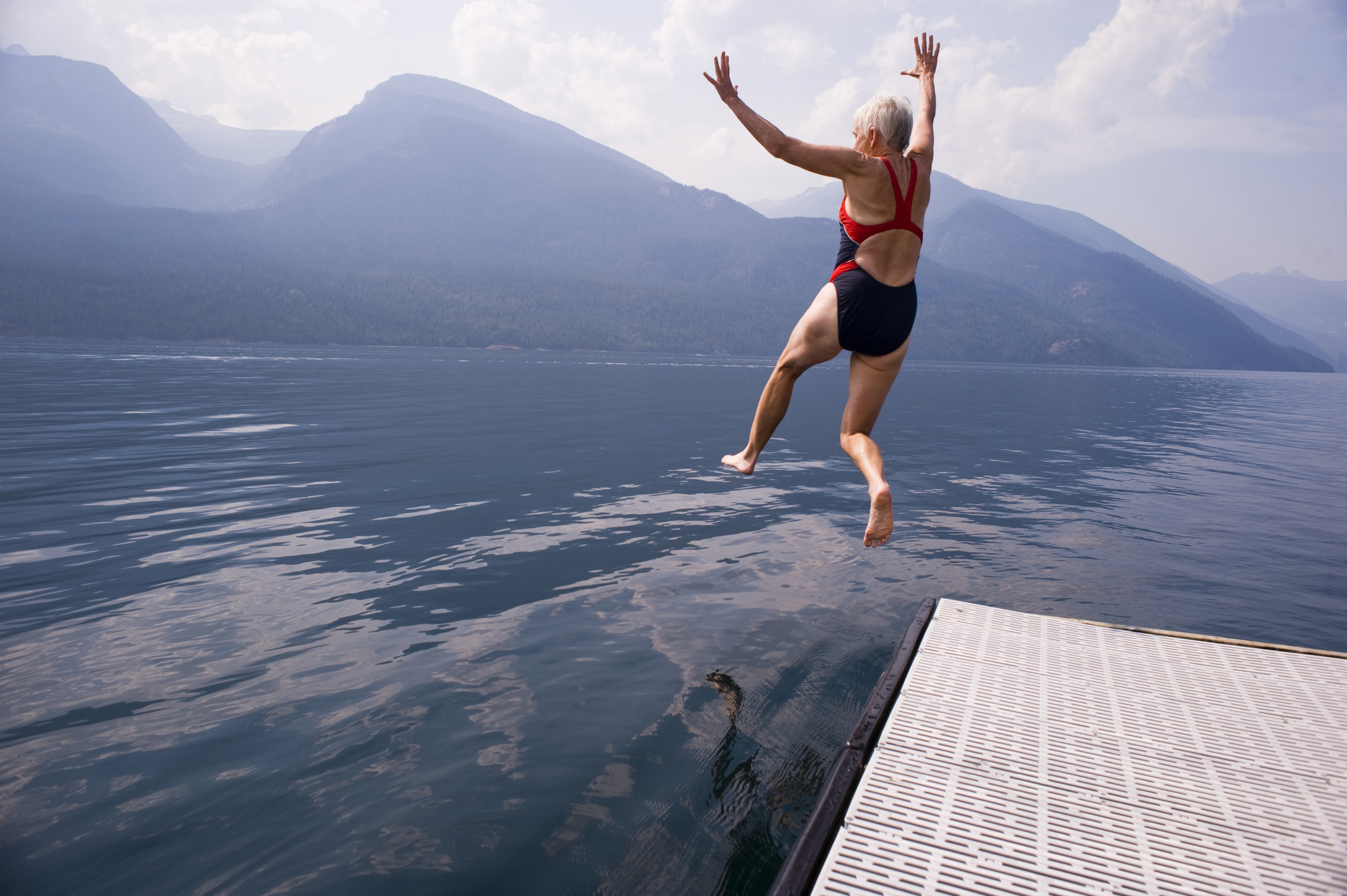 Person in a swimsuit joyfully jumps off a dock into a lake, with mountains visible in the background