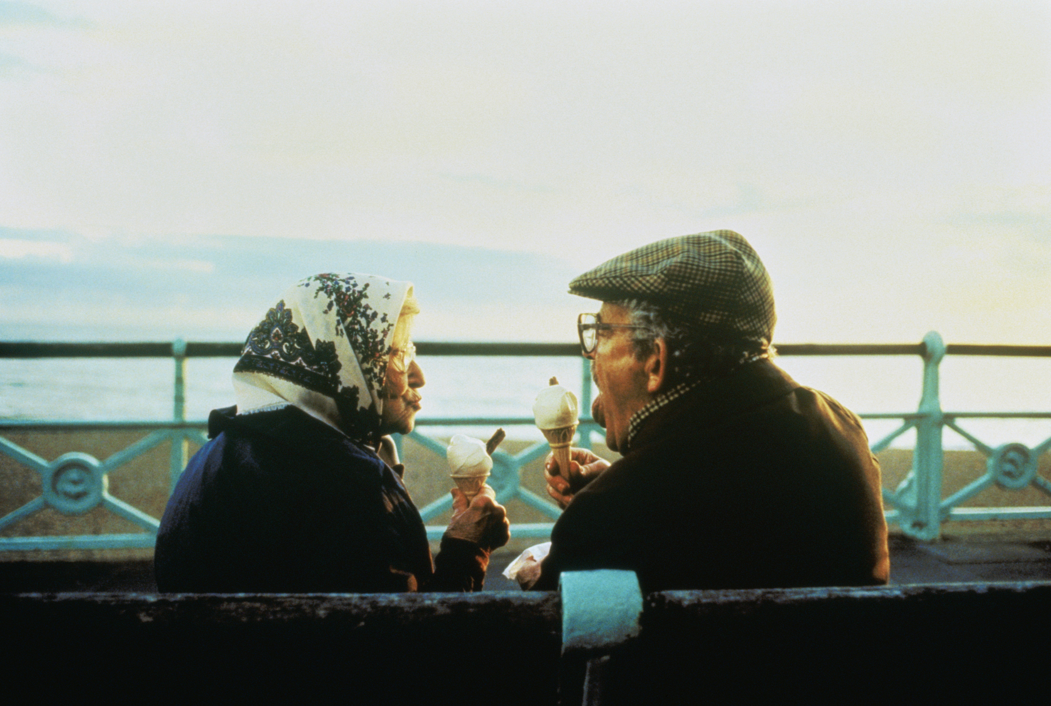 An elderly couple sits on a bench by the seaside, sharing ice cream and engaging in conversation