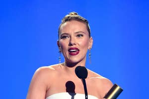 Scarlett Johansson speaks at an event, holding an award and standing in front of a blue background. She is wearing a strapless dress and gold earrings