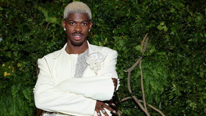 Lil Nas X posing confidently in an elegant, embellished white suit in front of lush greenery