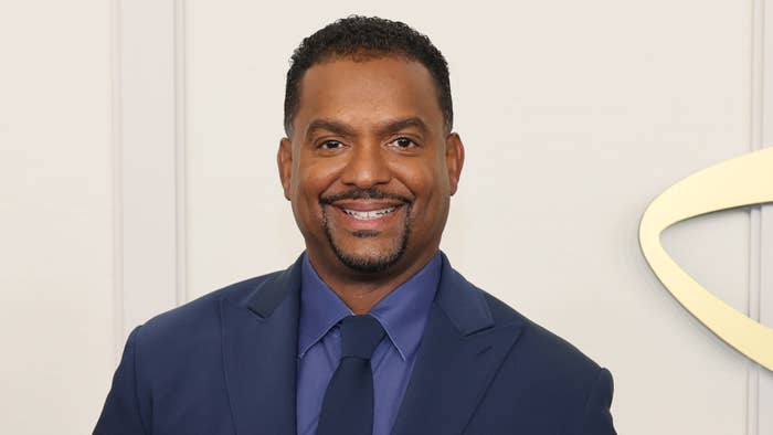 Alfonso Ribeiro smiling in a suit with a tie, standing against a plain background