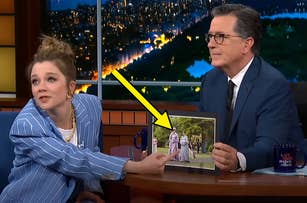 Actress Millicent Simmonds in a pinstripe suit points at a framed photo held by Stephen Colbert in a suit on a late-night show