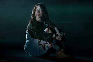 Billie Eilish sits on the ground in casual attire, including a graphic shirt and camouflage jacket, looking slightly to the side