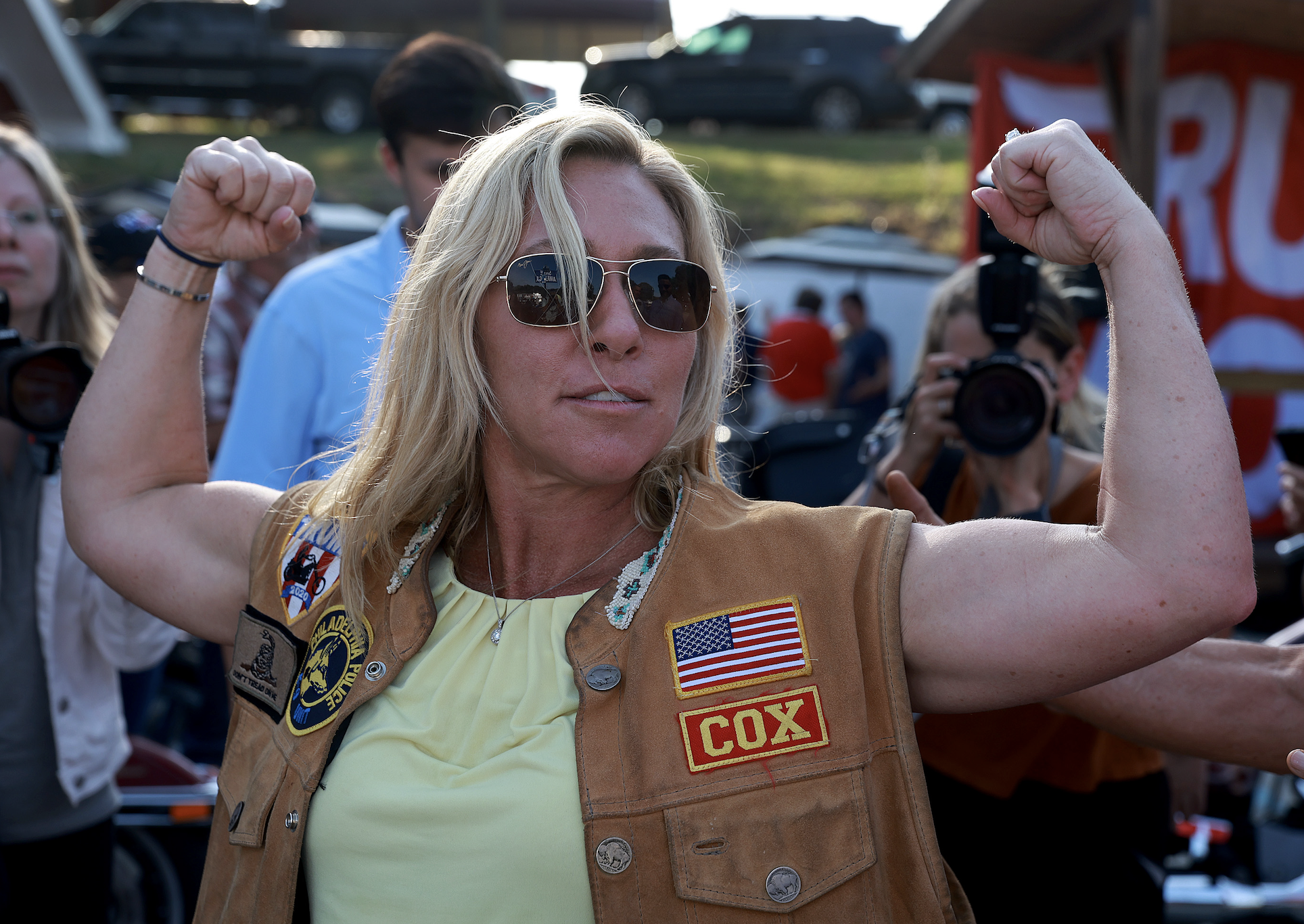 Marjorie Taylor Greene flexes her arms at an outdoor event, wearing a light-colored shirt and a vest with patches, including an American flag and "Cox". Photographers and people are in the background