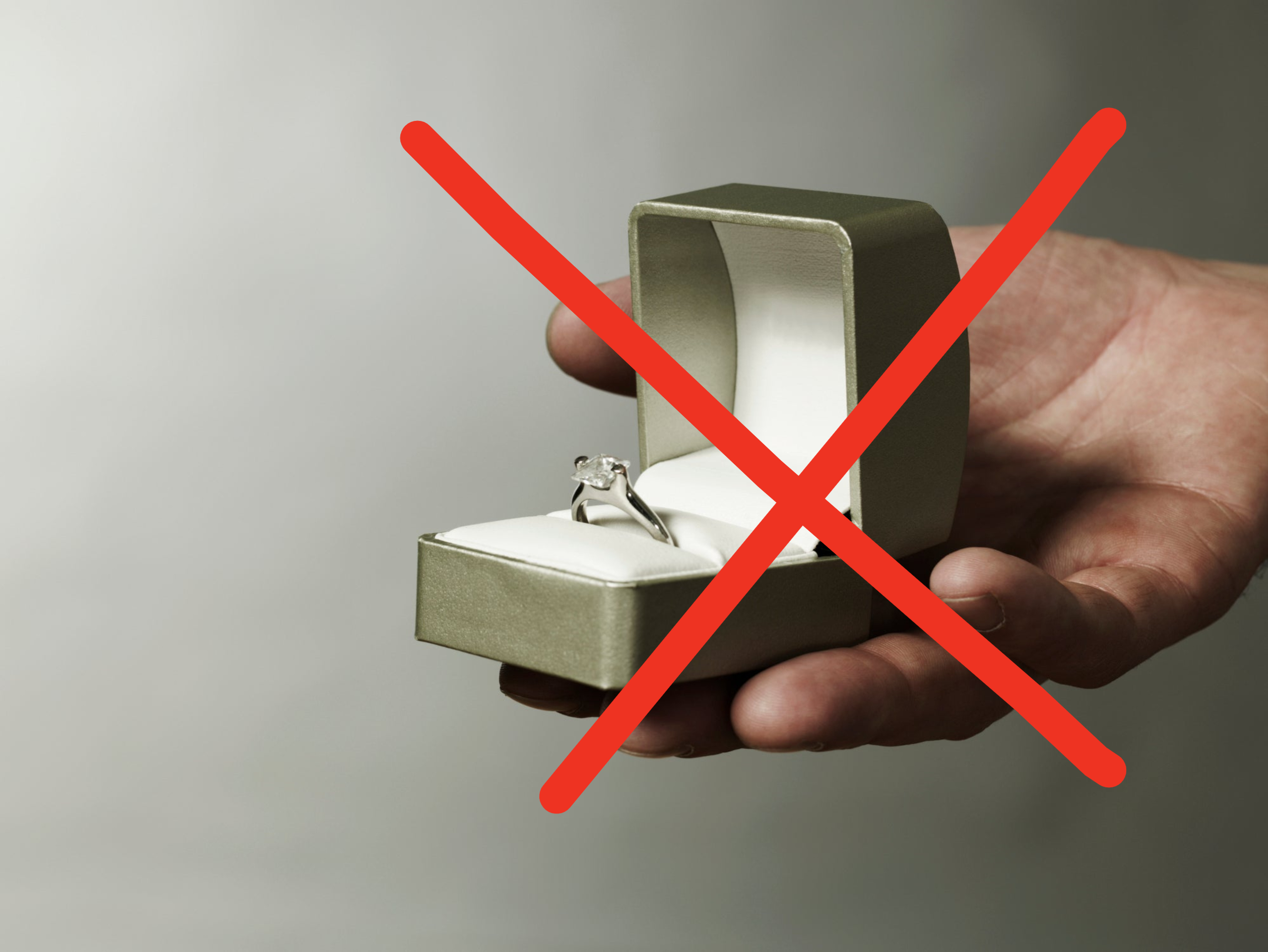 A hand holding an open ring box with an engagement ring inside with an X drawn over it