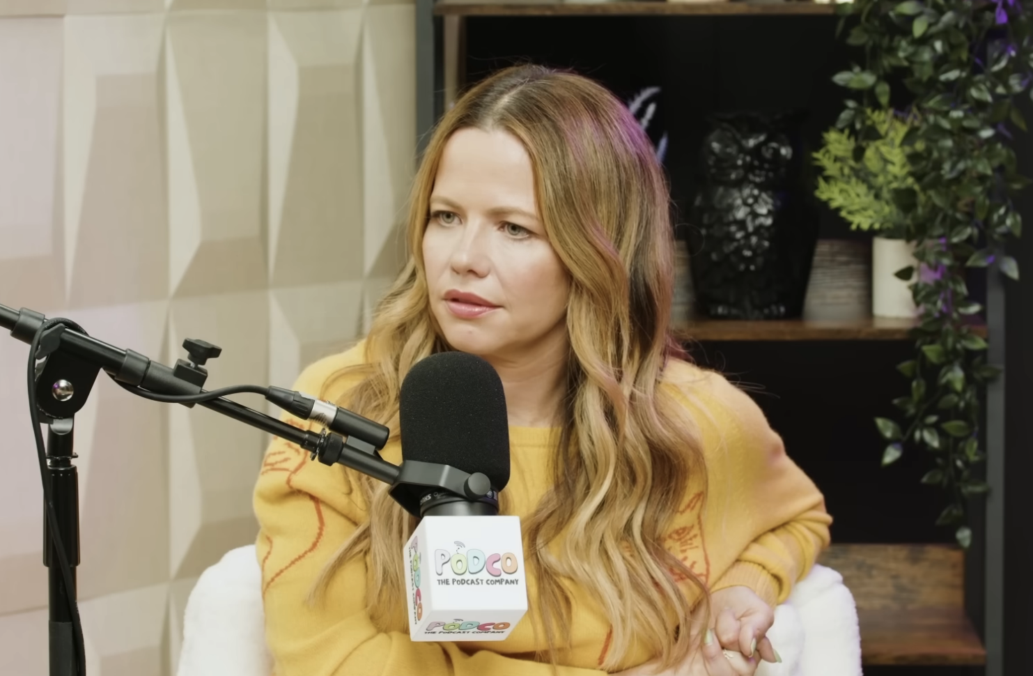Sasha Pieterse speaks into a microphone during an interview on Pretty Little Liars: True Crime YouTube show. She is wearing a long-sleeve top