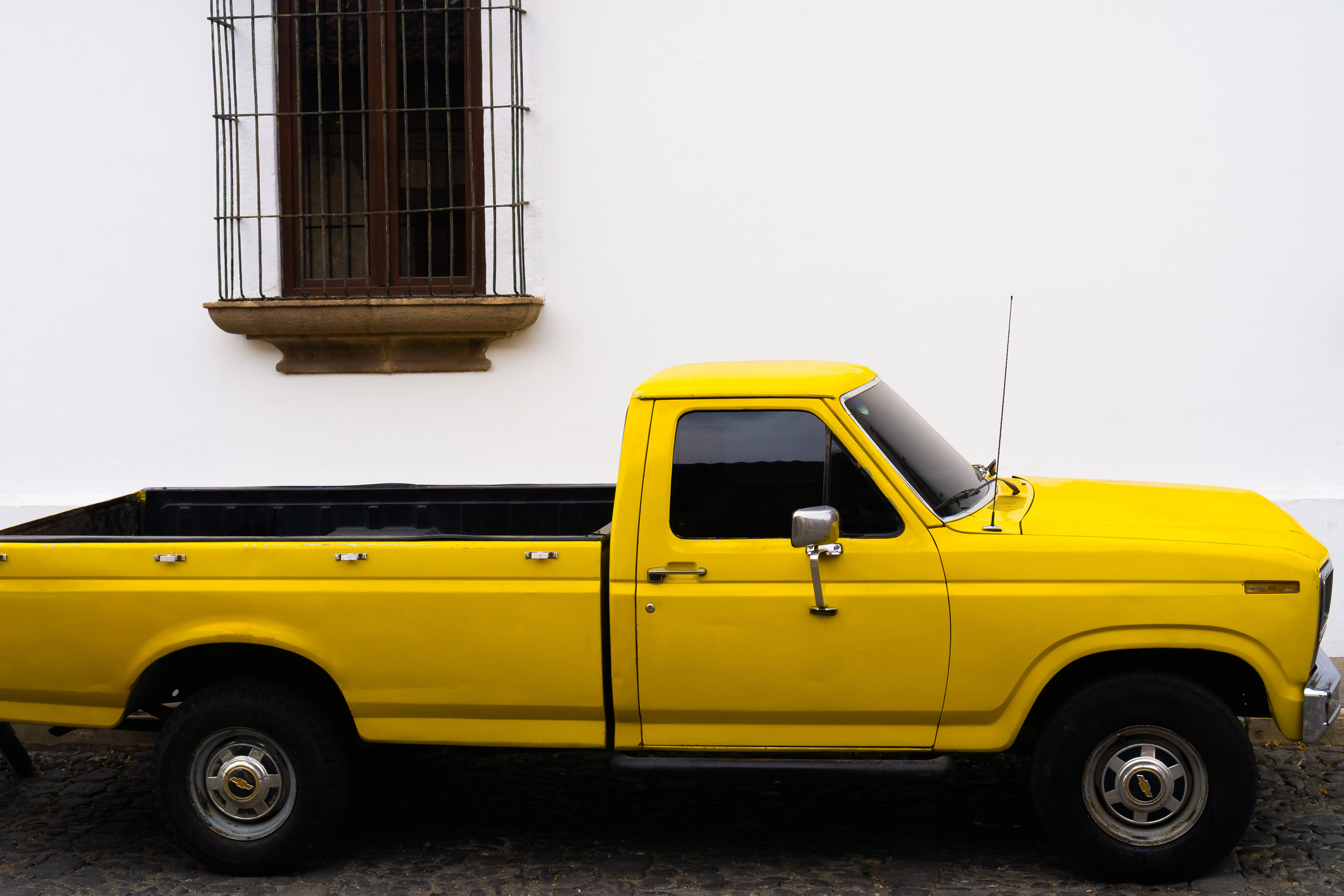 A yellow pickup truck is parked on a cobblestone street next to a white wall with a window