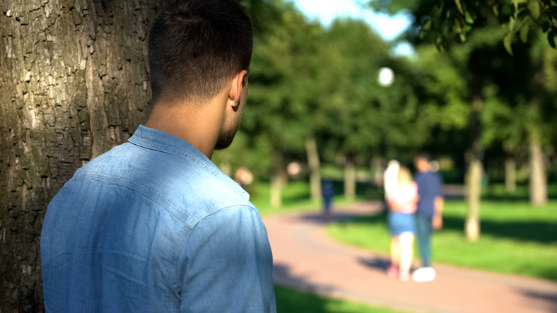 A man peers from behind a tree, watching a couple walking together in a park