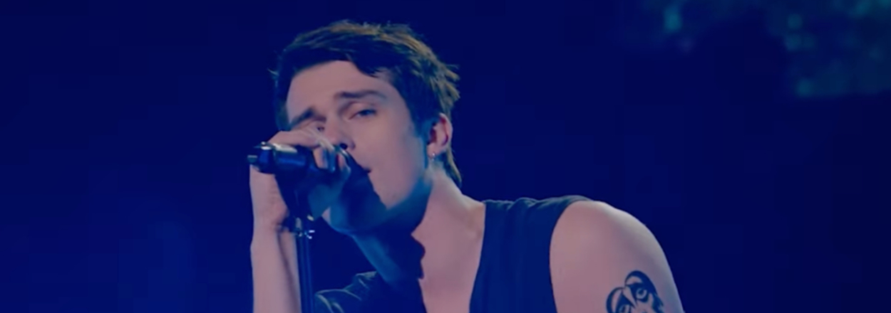 Nicholas Galitzine holding a microphone passionately performing on stage, wearing a sleeveless shirt, displaying tattoos on both arms