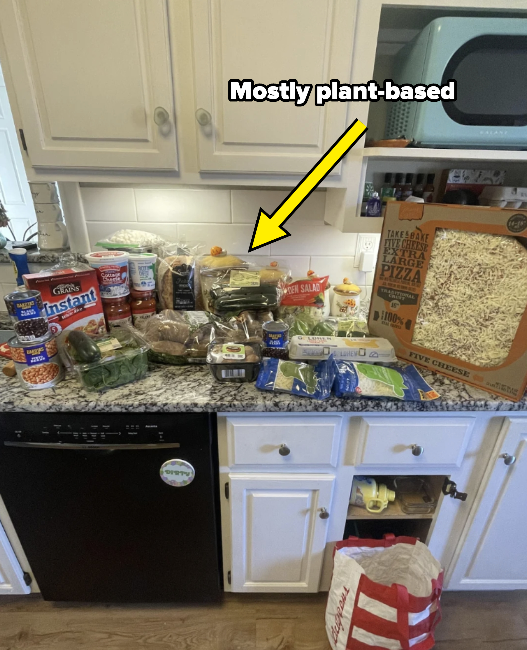 A kitchen counter with various grocery items displayed, including canned goods, vegetables, potato chips, drinks, and a large pizza. A red and white shopping bag is on the floor