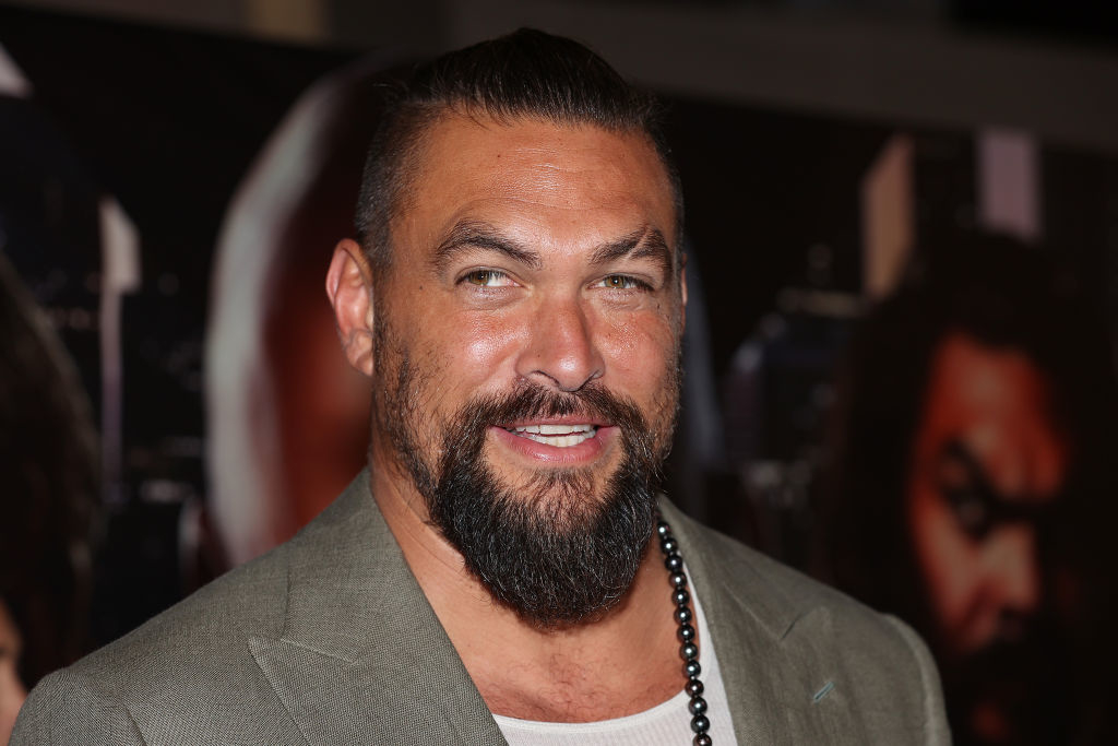 Jason Momoa smiling, wearing a blazer over a white shirt with a beaded necklace, at an event