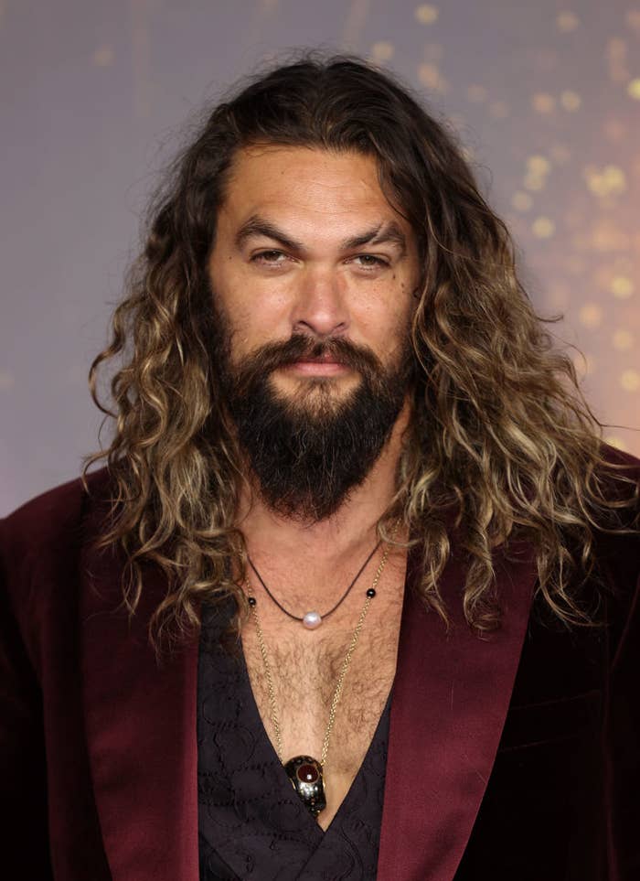 Jason Momoa in a dark velvet suit jacket over a dark shirt, accessorized with layered necklaces, at a formal event