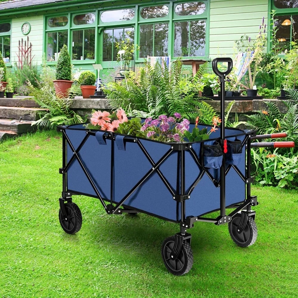 Gerobak taman yang dapat dilipat berisi berbagai macam bunga dan peralatan berkebun, diletakkan di halaman rumput depan rumah dengan jendela besar dan beberapa tanaman pot