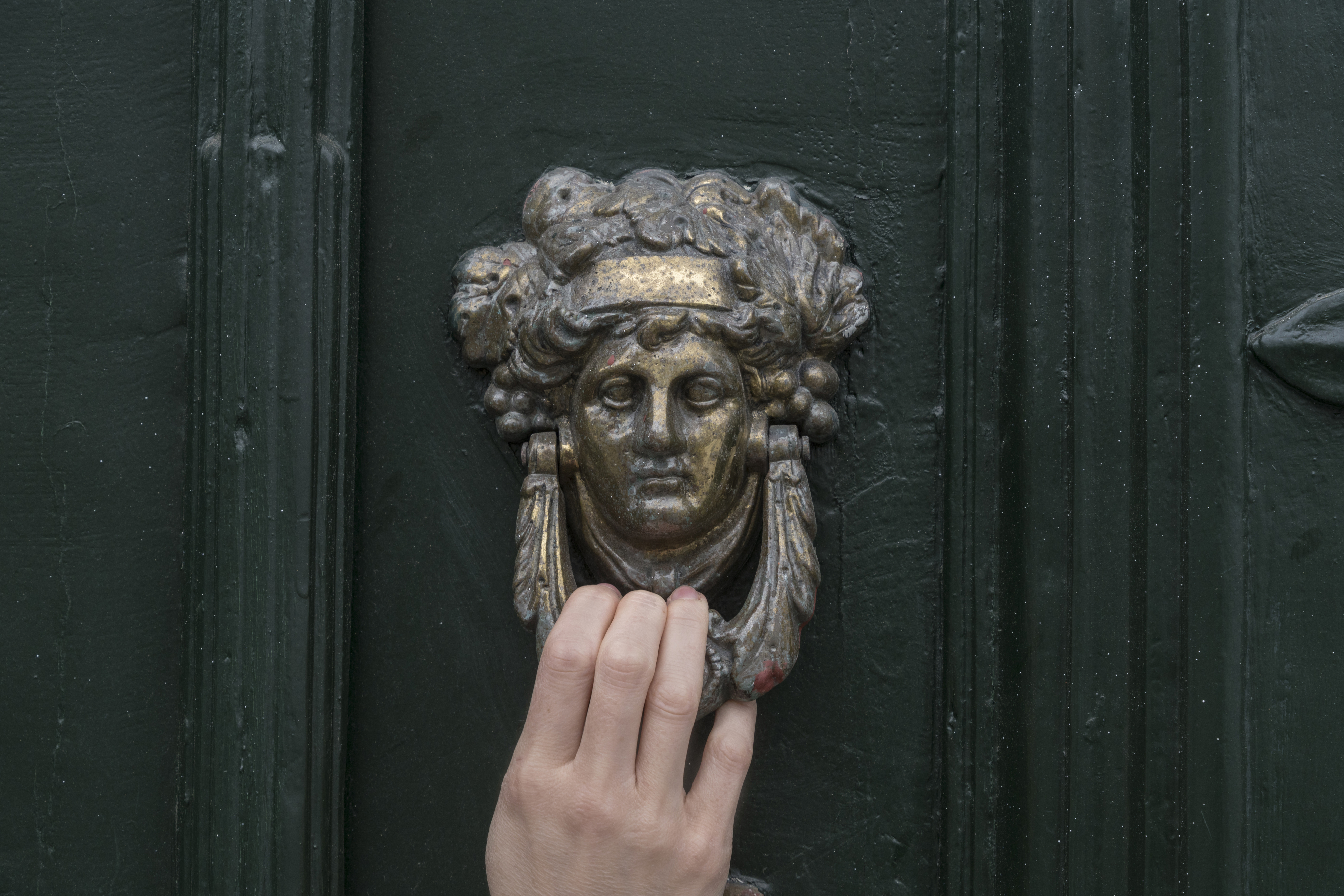 A hand is knocking on an old green door with a vintage brass door knocker shaped like a man's face
