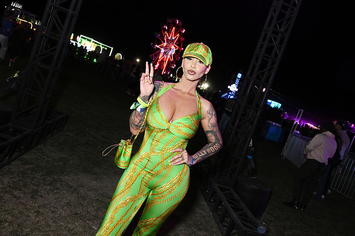 A person wearing a fitted, patterned jumpsuit and matching cap poses on a festival grounds at night, holding up a peace sign with their fingers