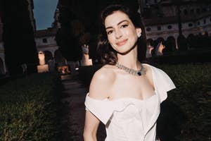 Anne Hathaway wearing an off-shoulder white dress with a statement necklace, smiling outdoors in an elegant garden setting