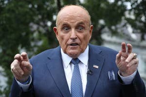 Rudy Giuliani speaking outdoors, dressed in a suit, raising both hands with his fingers in quotation mark gestures