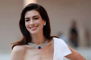 Anne Hathaway smiling, wearing a chic, sleeveless white outfit and a statement necklace, posing for the camera