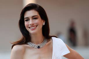 Anne Hathaway smiling, wearing a chic, sleeveless white outfit and a statement necklace, posing for the camera