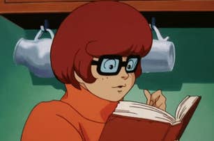 Animated character Velma Dinkley from Scooby-Doo is reading a book carefully while flipping pages. She wears her iconic glasses and orange turtleneck