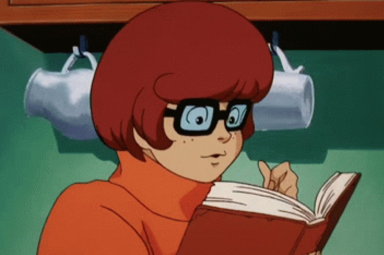 Animated character Velma Dinkley from Scooby-Doo is reading a book carefully while flipping pages. She wears her iconic glasses and orange turtleneck