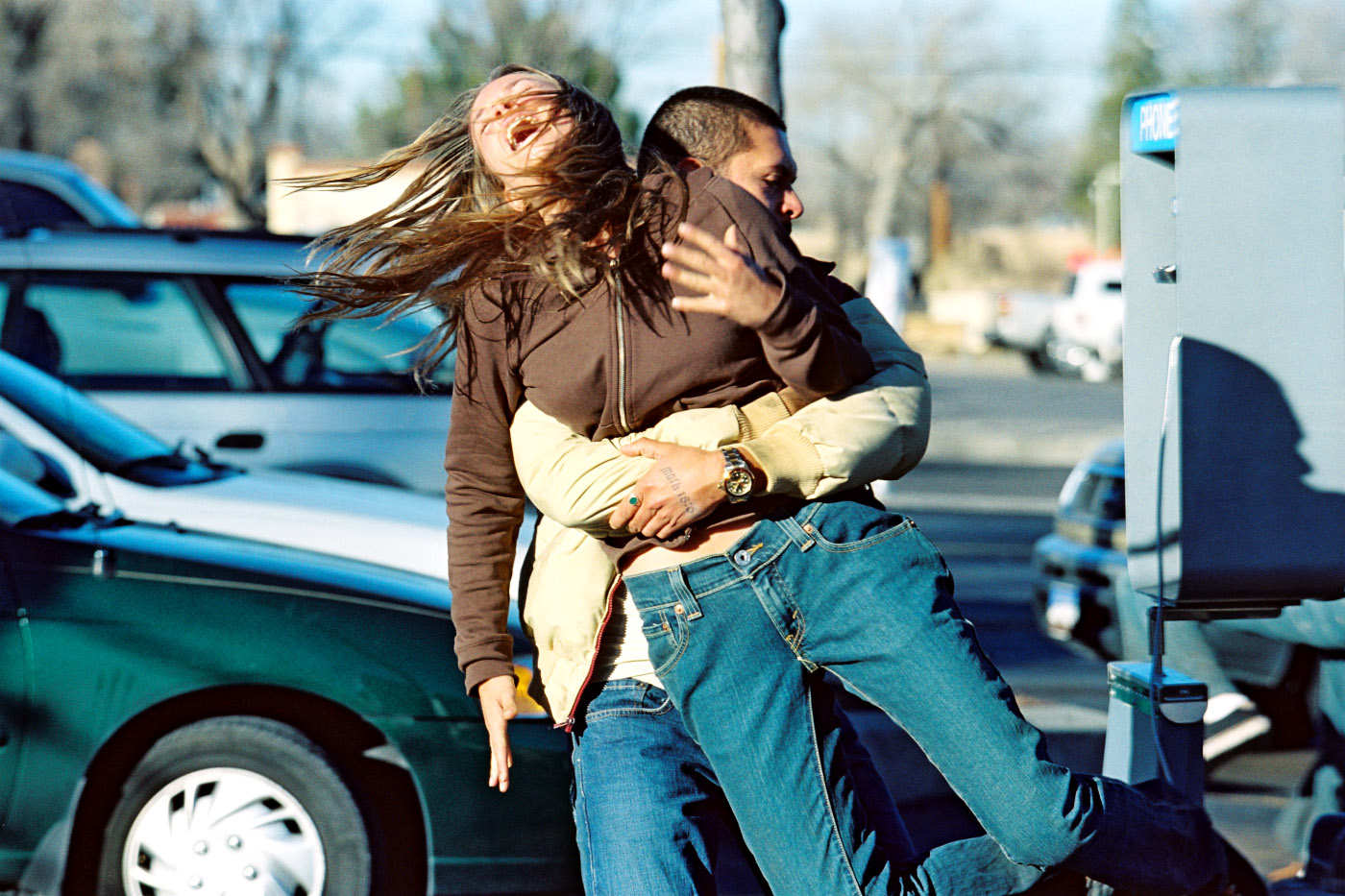 Two people are playfully struggling in a parking lot; one is lifting the other off the ground. Their expressions seem to convey excitement