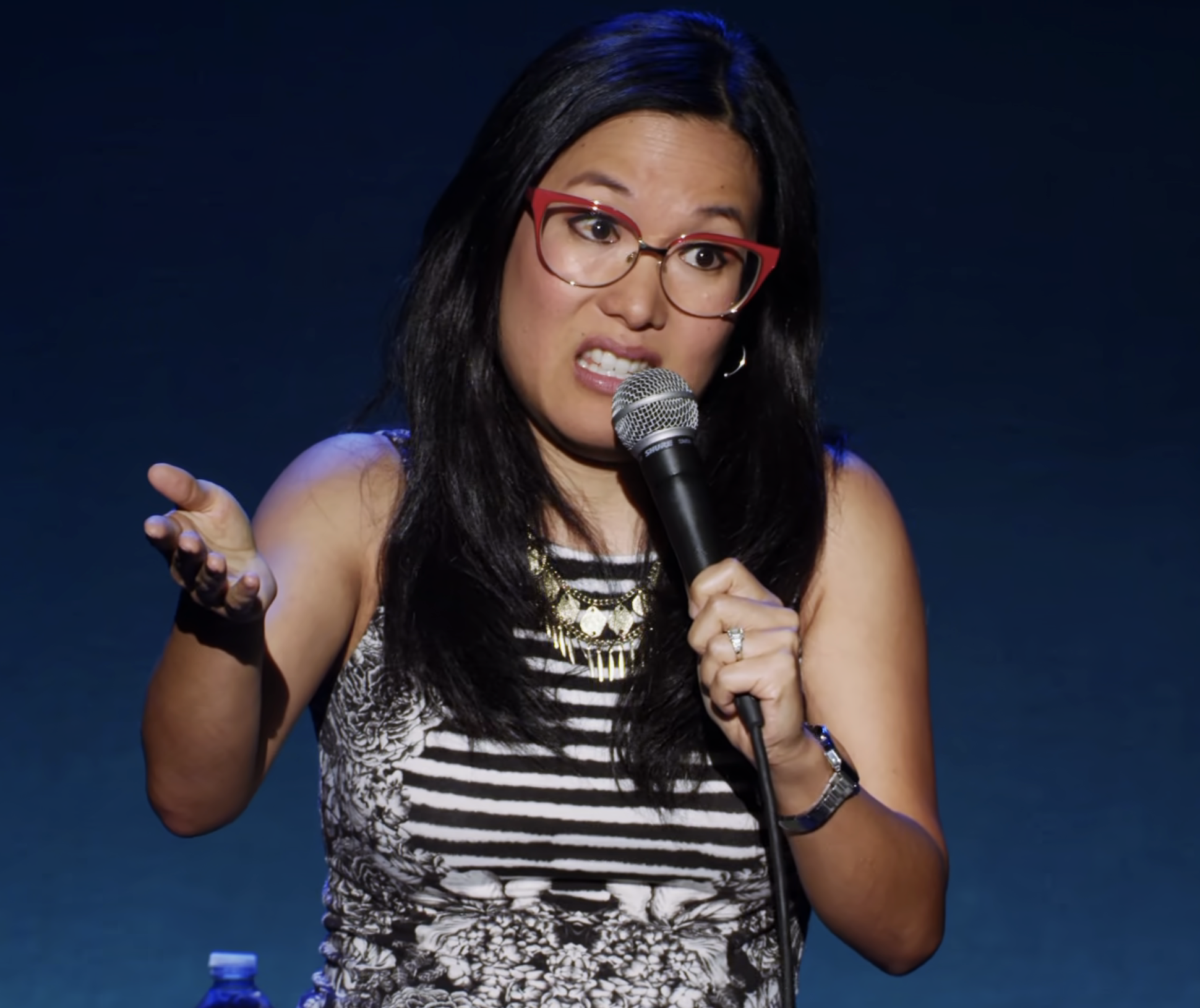 Ali Wong stands on stage, holding a microphone and gesturing with one hand. She is wearing a sleeveless patterned dress and glasses