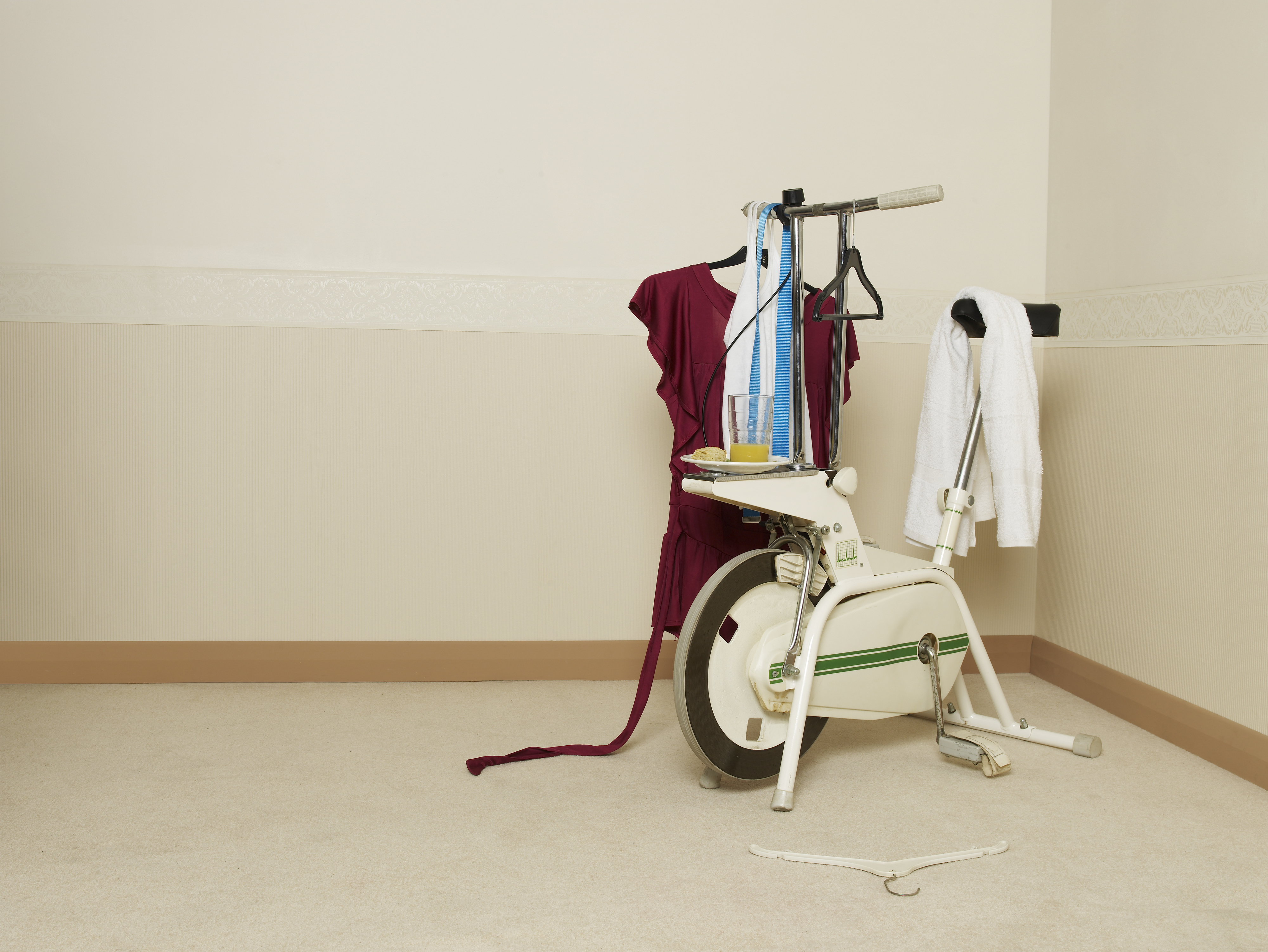 An exercise bike in a room serves as a makeshift clothes rack, holding a dress, towel, and other clothing items