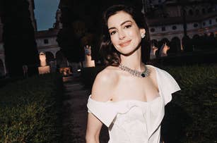 Anne Hathaway wearing an off-shoulder white dress with a statement necklace, smiling outdoors in an elegant garden setting