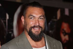 Jason Momoa at a public event, wearing a grey jacket over a white shirt and a bead necklace, smiling at the camera