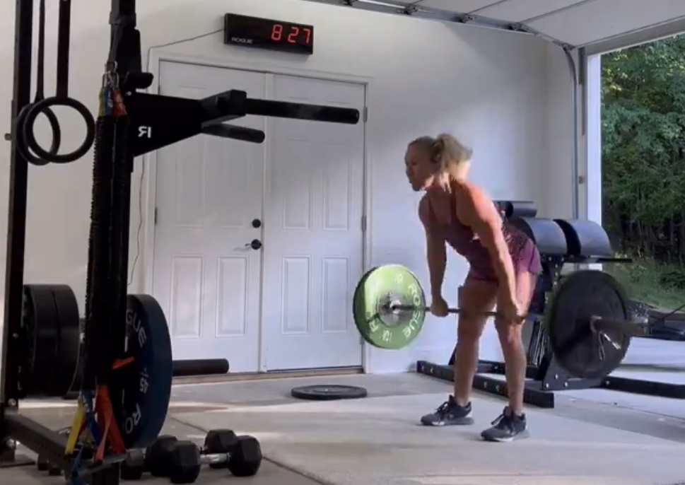 A person deadlifts a barbell in a home gym with various equipment. The wall clock reads 8:27