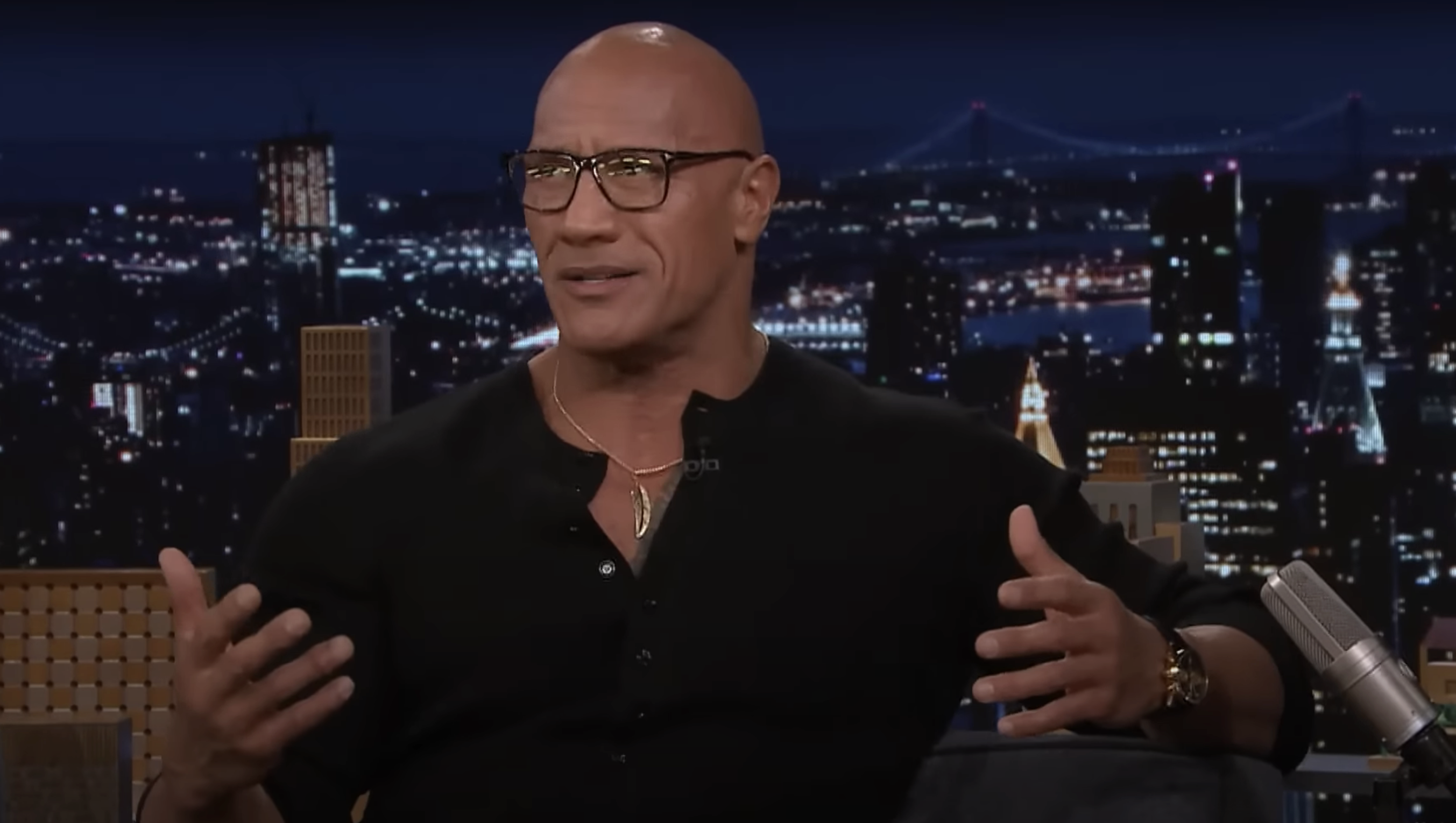 Dwayne Johnson, wearing glasses and a shirt, gestures while speaking on a nighttime talk show set with a cityscape backdrop
