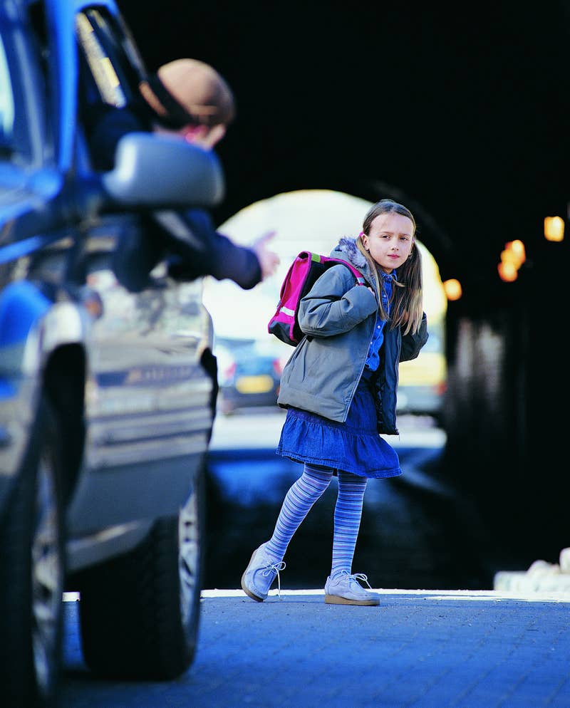 A girl with a pink backpack, wearing a blue dress and striped leggings, looks hesitant near a car with a person gesturing from the driver&#x27;s seat