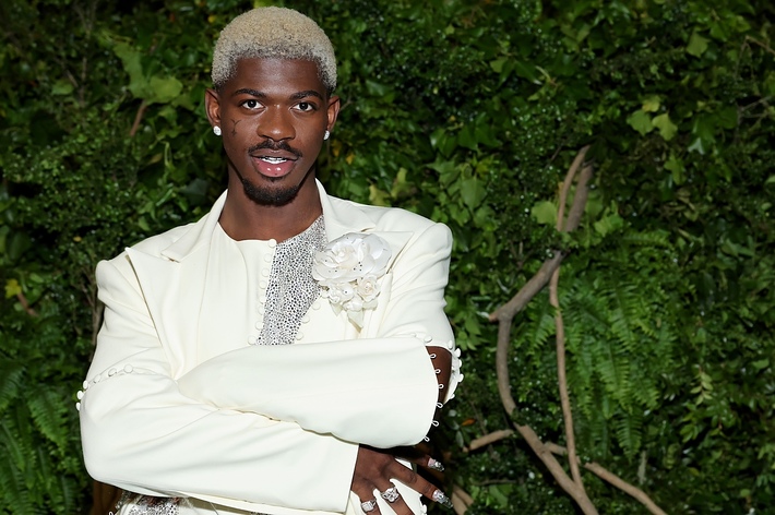 Lil Nas X poses outdoors wearing a cream-colored suit with a detailed floral brooch and sparkling glam jewelry. He stands against a backdrop of lush greenery
