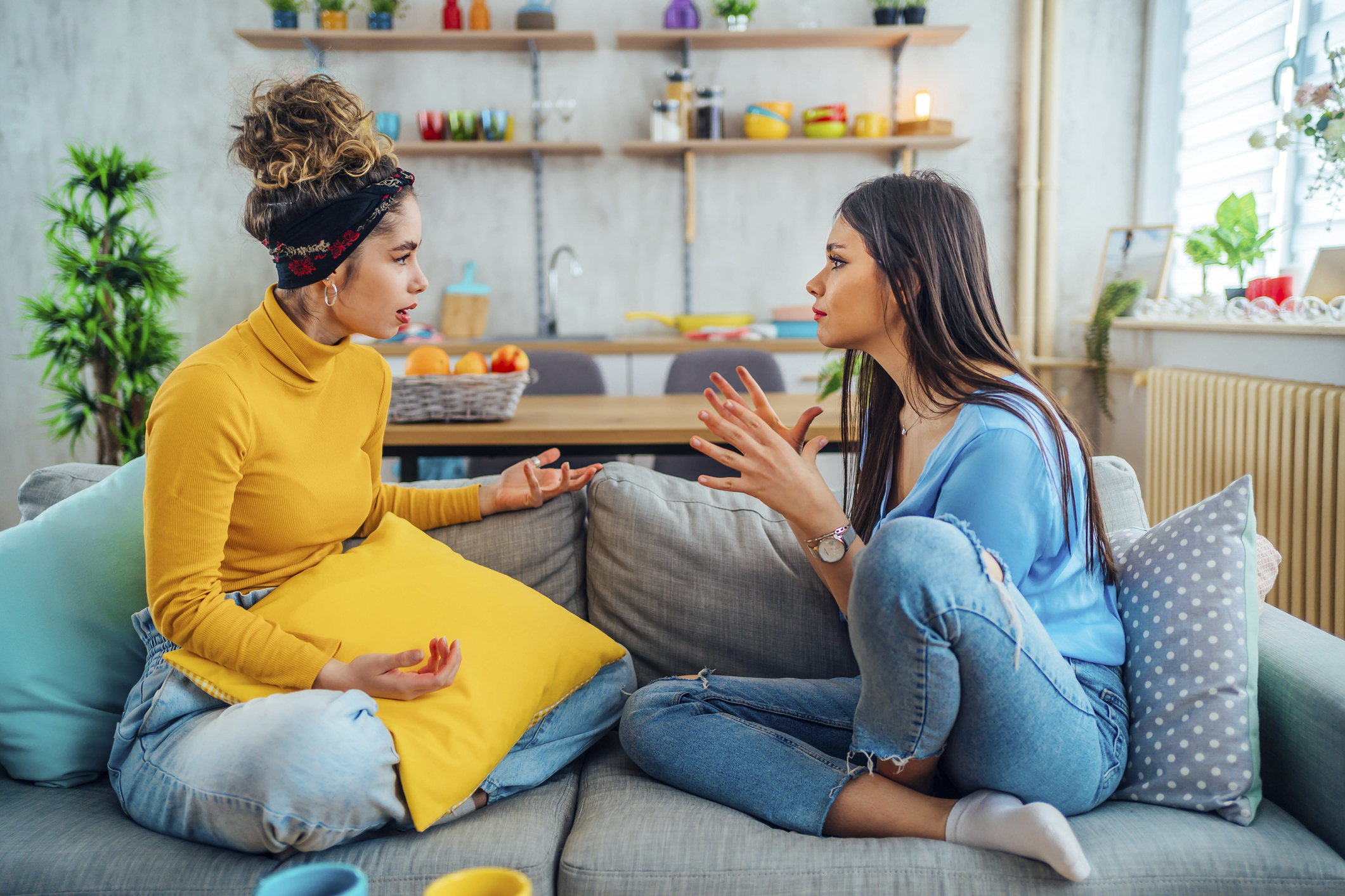 Two women sitting on a sofa having a serious conversation. One wears a yellow turtleneck and a headband, while the other wears a blue shirt