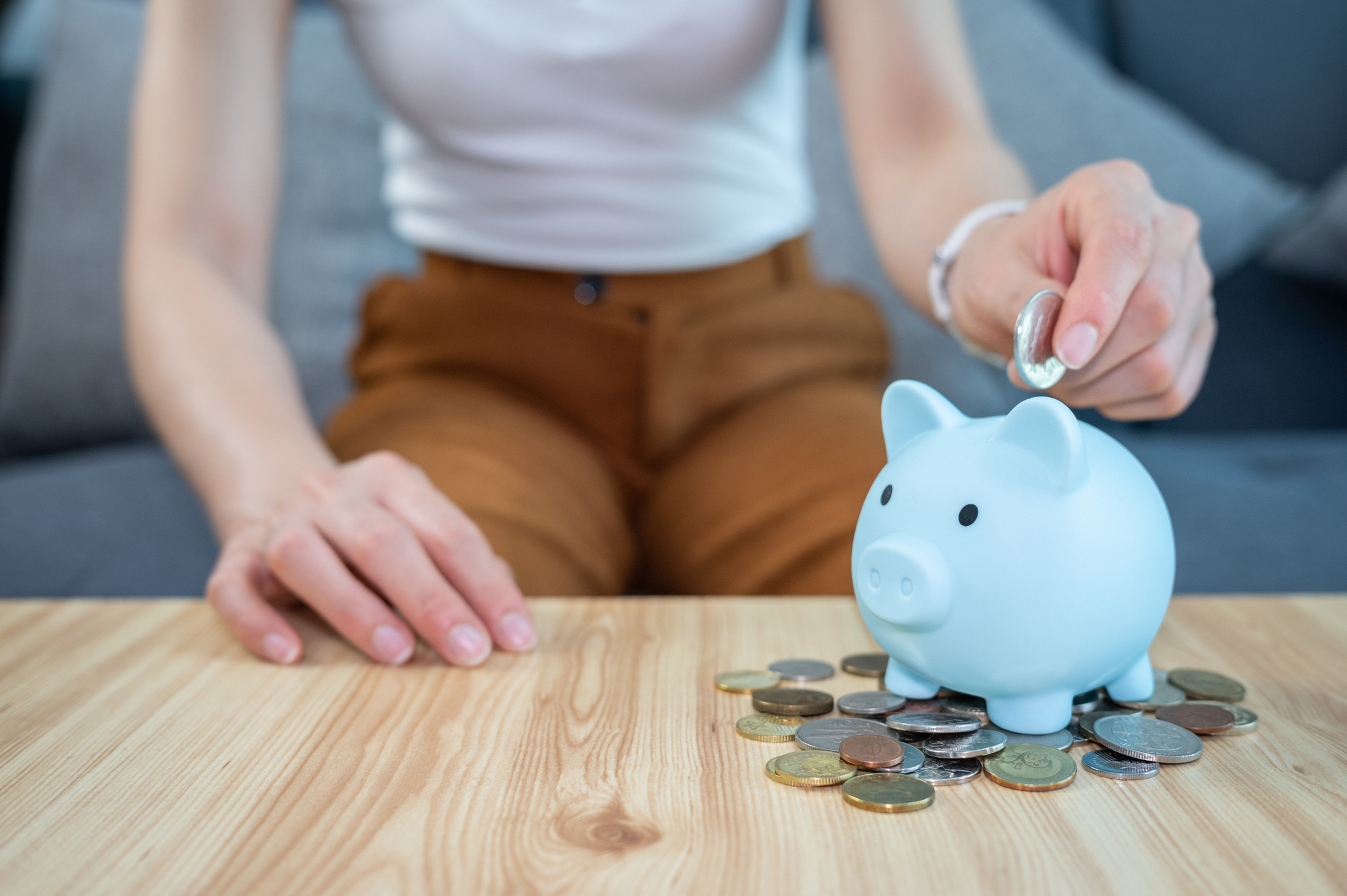A person places a coin into a blue piggy bank on a wooden table with scattered coins around it
