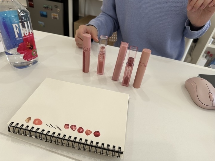 Person's hand near several lip gloss tubes and swatches of different lip gloss shades on a notebook, beside a water bottle and a computer mouse