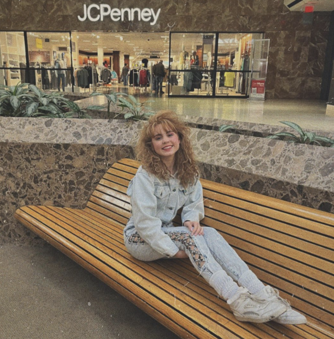 Ariana Greenblatt sits on a wooden bench in a mall, smiling in front of a JCPenney store, wearing a denim jacket, patterned pants, and white sneakers