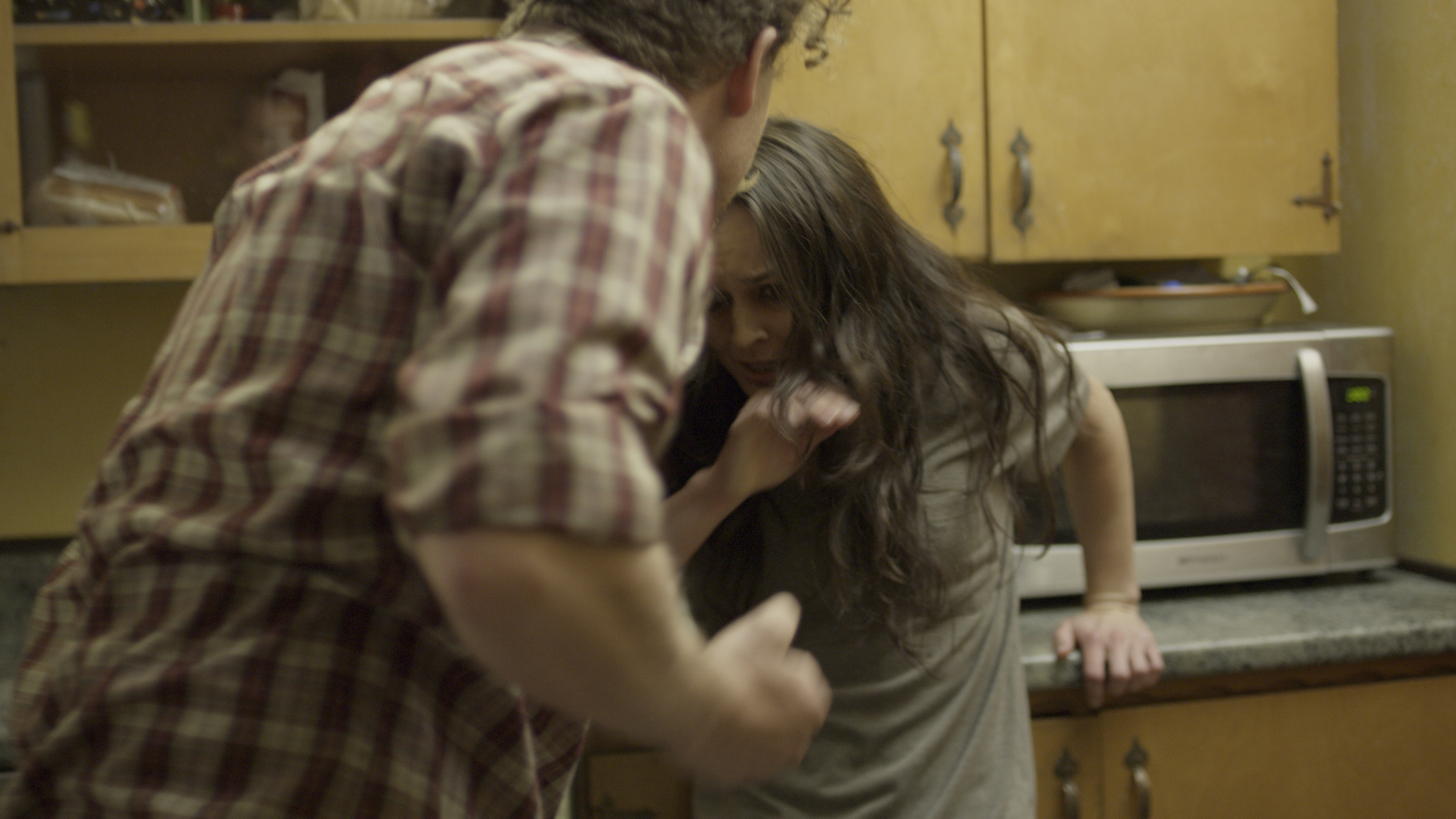 A man in a plaid shirt appears to be yelling at a frightened woman in a kitchen, with the woman cowering near a microwave