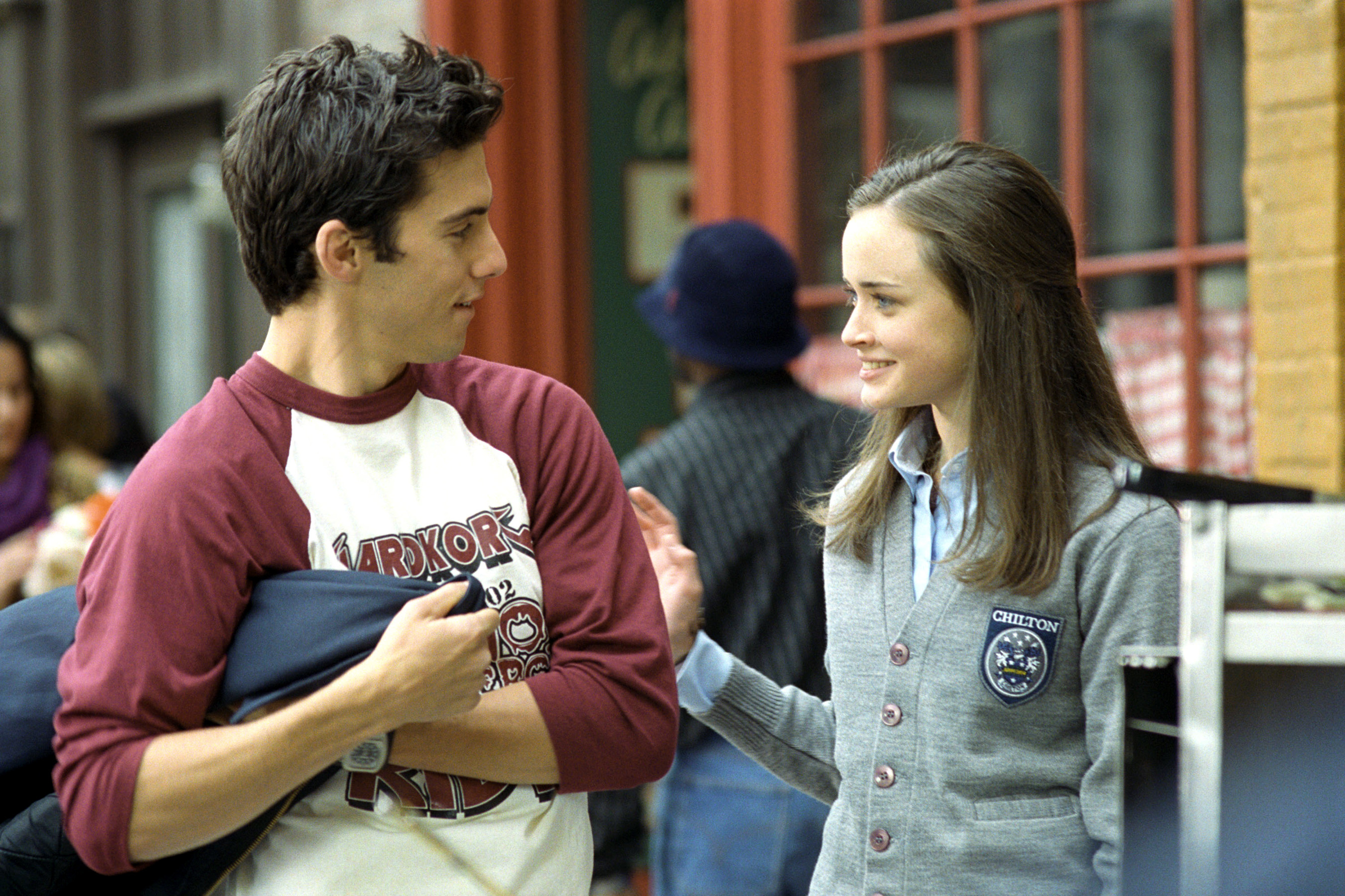 Milo Ventimiglia and Alexis Bledel on a street scene from "Gilmore Girls." Milo is wearing a casual shirt, and Alexis is in a school uniform with a Crest insignia
