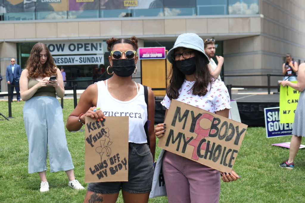 Two women at an outdoor protest holding signs that read &quot;Keep Your Laws Off My Body&quot; and &quot;My Body My Choice.&quot; Other people are visible in the background