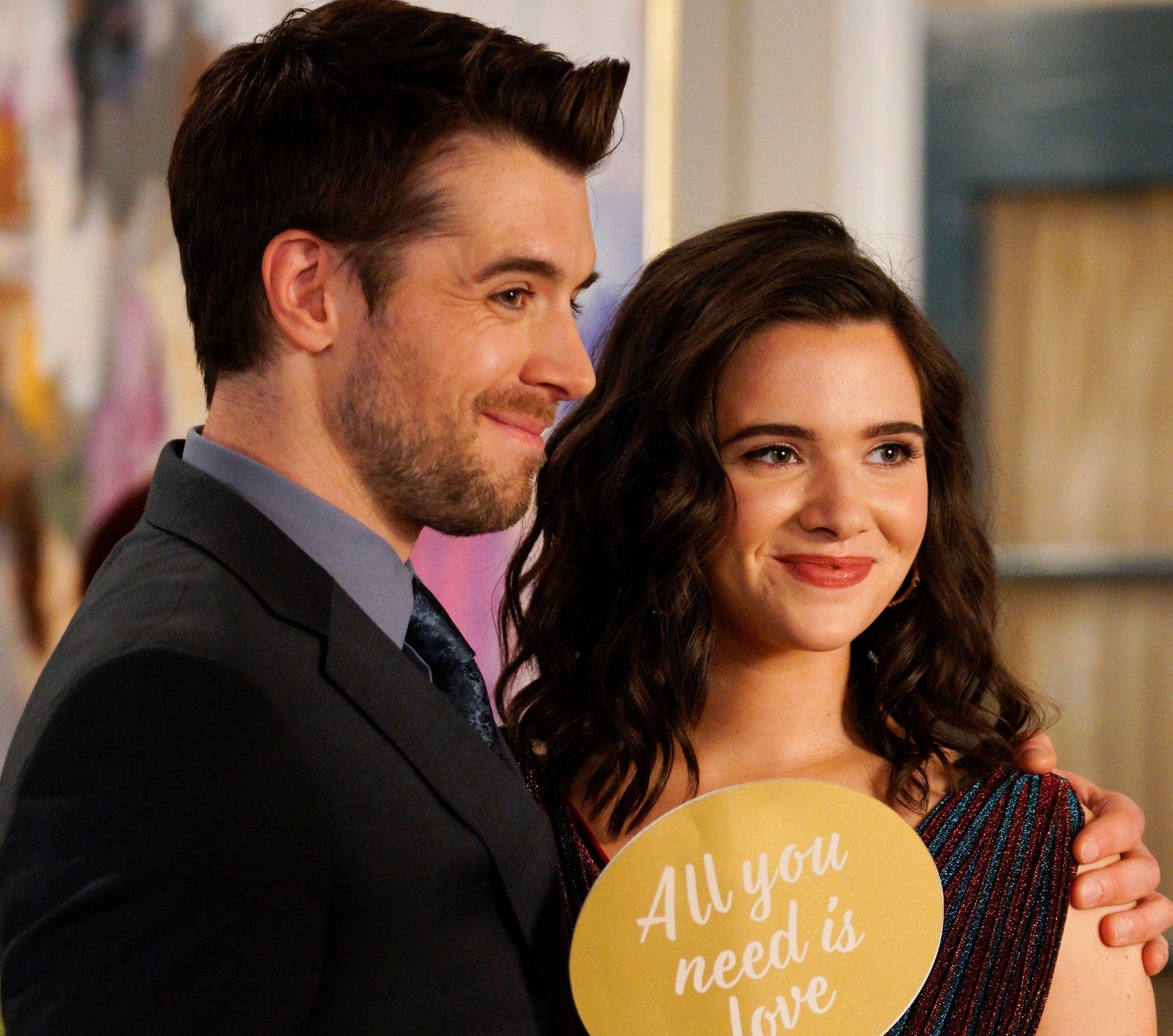 Two people smiling, the woman holds a sign reading "All you need is love." Both dressed semi-formal; the man in a suit and the woman in a sleeveless dress with sparkles